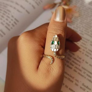 Puede incluir: Un anillo de serpiente dorado con un diseño de cabeza blanca y verde. El anillo se enrolla alrededor del dedo, con la cabeza de la serpiente en la parte superior y una cola curva. El anillo se lleva en una mano.