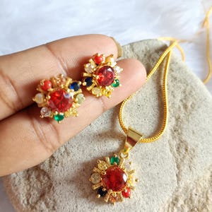 Peut inclure: Ensemble de bijoux dorés comprenant un collier pendentif et des boucles d'oreilles assorties. Chaque pièce présente une pierre rouge centrale entourée de petites pierres précieuses colorées. Les boucles d'oreilles sont de type clous, et le collier a une chaîne délicate.