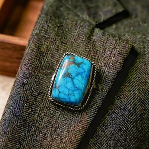 May include: A rectangular turquoise ring with a silver rope-like border. The turquoise stone has a vibrant blue color with brown veining. The ring is set against a dark green herringbone fabric.