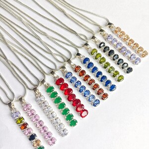 May include: A selection of silver necklaces, each with a pendant adorned with a line of colourful gemstones. The gemstones are in shades of pink, green, red, blue, and orange. The necklaces are presented on a white surface.
