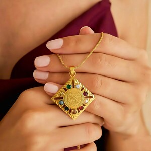 Könnte beinhalten: Eine goldfarbene Anhängerkette mit einem quadratischen, rautenförmigen Anhänger. Der Anhänger hat ein zentrales kreisförmiges Design, das von bunten Edelsteinen und einem strukturierten Muster umgeben ist. Die Halskette wird von einer Person gehalten.