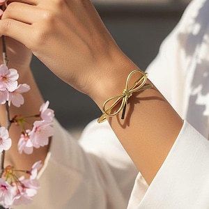 Puede incluir: Una pulsera dorada con forma de lazo se lleva en la muñeca. La pulsera tiene un diseño delicado, con el lazo como punto focal. El fondo está borroso, con un toque de flores rosas y una camisa blanca.