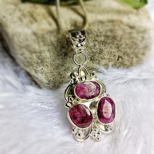 Puede incluir: Un colgante de plata con tres piedras preciosas ovaladas de color rojo intenso. El colgante tiene un diseño ornamentado con remolinos decorativos y un lazo para una cadena. El fondo es una vista borrosa de una piedra y tela blanca.