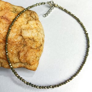 Op de afbeelding: Een delicate ketting met kleine, facetgeslepen kralen in goud- en olijfgroene tinten. De ketting is geregen op een dunne, zilverkleurige ketting en heeft een verstelbare sluiting. De ketting wordt getoond op een getextureerde, bruinachtige steen.