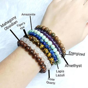 May include: A collection of beaded bracelets in various colors and gemstones, including mahogany obsidian, tiger's eye, rose quartz, and amethyst. The bracelets are displayed on a wrist, showcasing the different textures and hues of the natural stones. The word "Energized" is written on the image.