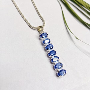 May include: A silver necklace with a pendant of seven oval, blue gemstones. The pendant hangs from a fine silver chain. The gemstones are arranged vertically, creating a striking design. The background is white with green foliage.