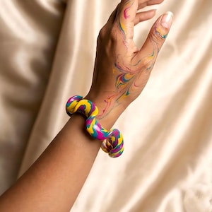 Puede incluir: Una colorida pulsera ondulada en amarillo, rosa y turquesa se lleva en la muñeca. El brazo está contra un fondo color crema. La mano y el brazo tienen trazos de pintura coloridos.
