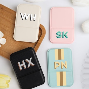 May include: Four rectangular phone cases in cream, pink, black, and light blue, each with a different set of initials. The cream case has "WH", the pink case has "SK", the black case has "HX", and the light blue case has "PN".