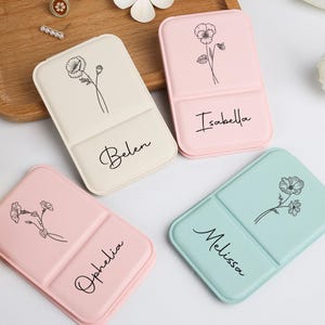 Könnte beinhalten: Vier personalisierte, zweifach gefaltete Kosmetikkoffer in Pastellfarben: Creme, Rosa und Mintgrün. Jeder Koffer hat eine florale Strichzeichnung und einen Namen in Schreibschrift: Belen, Isabella, Ophelia und Melissa. Die Koffer sind auf einem Holztablett angeordnet.