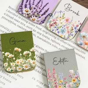Może przedstawiać: Pięć kolorowych skórzanych zakładek do książek z kwiatowymi wzorami i imionami: "Adora", "Brook", "Coral", "Gina" i "Edith". Każda zakładka ma inny układ kwiatowy i kolor tła, w tym lawendę, dzikie kwiaty i maki. Zakładki są umieszczone na otwartej książce.