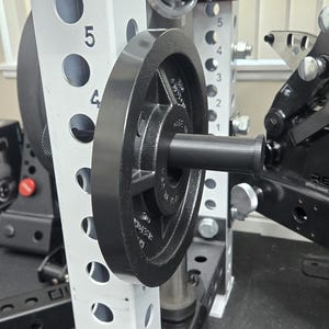 May include: A black weight plate attached to a weight machine. The weight plate is round with a central hole and a handle. The machine has a silver frame with numbered holes and black components.