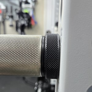 May include: Close-up of a weightlifting bar handle with a textured silver grip and a black, knurled end cap. The bar is attached to a white weight rack, suggesting a gym or fitness equipment setting.