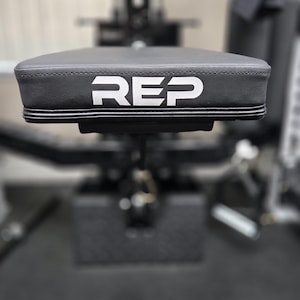 May include: A black weight bench seat with the white letters "REP" printed on the top. The seat is made of a textured material and is part of a larger weight training machine. The background is blurred.