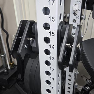 May include: Close-up of a weightlifting rack with black attachments and a white frame. The frame has numbered holes for adjusting the attachments. The attachments are designed to hold weight plates for strength training exercises.