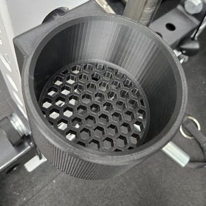 May include: A black, 3D-printed cup holder with a honeycomb pattern at the bottom. The cup holder is mounted on a weightlifting rack, designed to hold a beverage or other small items during workouts. The holder is made of textured plastic.