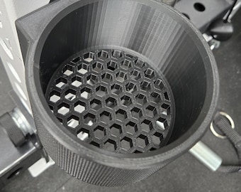 May include: A black, 3D-printed cup holder with a honeycomb pattern at the bottom. The cup holder is mounted on a weightlifting rack, designed to hold a beverage or other small items during workouts. The holder is made of textured plastic.