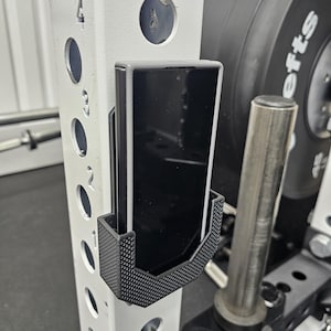 Pode incluir: Um telefone preto é segurado em um suporte de telefone preto e cinza preso a um rack de peso de metal branco. O suporte do telefone tem um padrão texturizado. Uma placa de peso é visível no fundo.