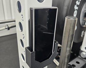 May include: A black phone is held in a black and gray phone holder attached to a white metal weight rack. The phone holder has a textured pattern. A weight plate is visible in the background.