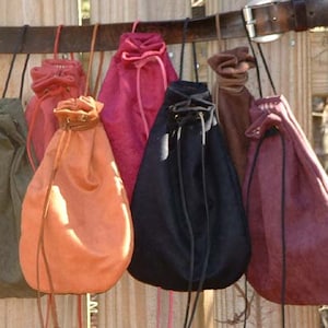 May include: Seven drawstring bags made of different fabrics and colors hanging on a wooden fence. The bags are all different sizes and shapes, and some have leather accents.
