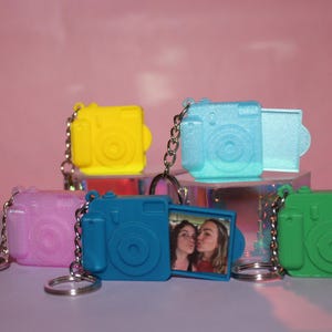 May include: A collection of colorful camera-shaped keychains. The keychains are in yellow, pink, blue, and green. One blue keychain has a small photo inside. Each keychain has a silver chain and ring.
