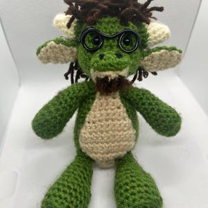 May include: A handmade green crochet doll with a tan belly and cream-colored accents. The doll has brown yarn hair, black wire-rimmed glasses, and a small brown beard. The doll's arms and legs are outstretched.