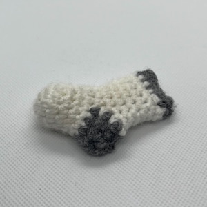 May include: A small, handmade crochet item in the shape of a sock. The sock is primarily white with gray accents on the heel and toe. The crochet stitches are visible, giving it a textured appearance. The item is likely a decorative piece or a small toy.