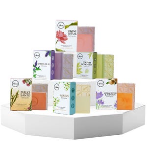 Könnte beinhalten: Eine Präsentation verschiedener Luxus-Badebars, jede in einer einzigartigen Schachtel. Die Schachteln sind weiß mit farbenfrohen botanischen Illustrationen und Text. Die Bars sind in den Farbtönen Rosa, Orange und Grün gehalten. Die Präsentation befindet sich auf einer weißen, geometrischen Plattform.