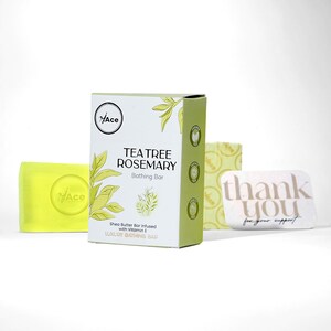 以下が含まれることがあります： ティーツリーとローズマリーのバスバーのディスプレイ。メイン商品は「Tea Tree Rosemary Bathing Bar」と書かれた白と緑の箱です。黄色の石鹸とサンキューカードも表示されています。