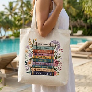 May include: A cream tote bag with a design of stacked books and floral elements. Each book spine has a title. The bag is held by a person in a white dress, with a tropical beach scene in the background.