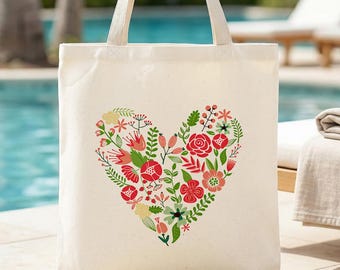Puede incluir: Bolso tote de color crema con un diseño de corazón floral. El corazón está lleno de flores rojas, rosas y amarillas, hojas verdes y bayas. El bolso tiene asas largas de color crema y está hecho de un material de lona resistente.