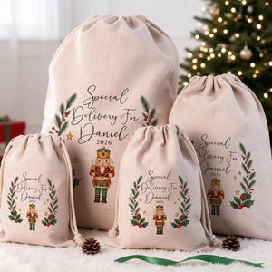 May include: Four beige canvas drawstring gift bags of varying sizes, each printed with the text Special Delivery For Daniel 2026, a nutcracker illustration, and a wreath of pine branches, red berries, and small gold stars.