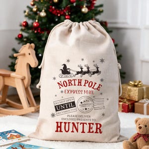 Può includere: Un sacco regalo in tela beige con coulisse, caratterizzato da una grafica nera di Babbo Natale su una slitta. Il testo recita NORTH POLE EXPRESS MAIL, DO NOT OPEN UNTIL DECEMBER 25TH e HUNTER, con icone di fiocchi di neve e timbri.