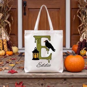 Peut inclure: Un sac fourre-tout en toile blanche orné d'une grande lettre E verte avec un effet dégoulinant. Un corbeau noir est perché sur la lettre et une lanterne noire à la lueur orange est posée à la base. Le nom Eddie est écrit en noir en dessous.