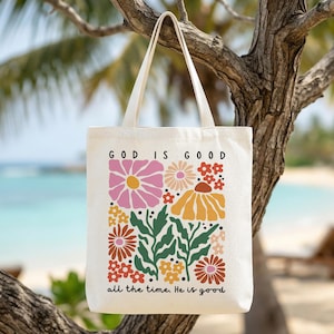 Könnte beinhalten: Cremefarbene Tragetasche mit floralem Design und dem Text "GOD IS GOOD all the time. He is good." Das Design zeigt rosa, orange und rote Blumen mit grünen Blättern. Die Tasche hängt an einem Ast.