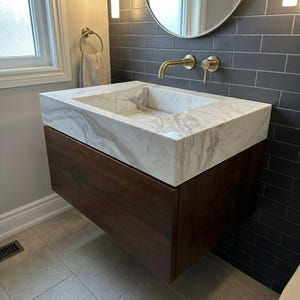 May include: A floating bathroom vanity featuring a dark brown wooden cabinet base and a thick white marble countertop with grey veining. The sink is integrated into the marble, complemented by a wall-mounted brushed gold faucet and handle.