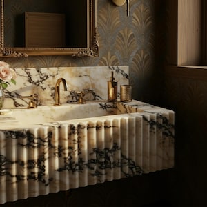 K&ouml;nnte beinhalten: Ein luxuri&ouml;ses Badezimmerwaschbecken aus Marmor mit goldenen Armaturen. Das Waschbecken hat ein geriffeltes Design und wird durch einen goldenen Seifenspender und Becher erg&auml;nzt. Der Marmor hat schwarze Aderungen.