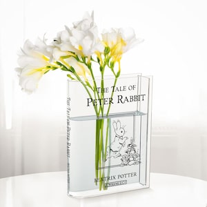 May include: A clear, book-shaped vase filled with water and white flowers. The vase features the title "The Tale of Peter Rabbit" and an illustration of Peter Rabbit. The vase is on a white surface.