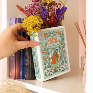 Könnte beinhalten: Eine buchförmige Keramikvase mit dem Titel "The Secret Garden" von Frances Hodgson Burnett, gefüllt mit bunten Trockenblumen. Das Vasendesign zeigt ein Mädchen in einem roten Mantel und grünen Kleid. Die Vase wird in der Nähe eines Bücherregals gehalten.