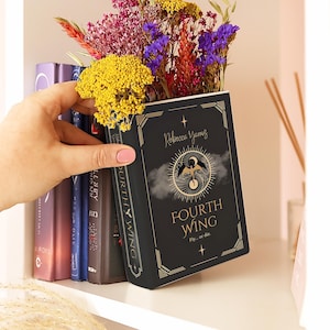 May include: A black book-shaped vase with gold detailing, featuring the text "FOURTH WING" and "Fly... or die". The vase is filled with vibrant flowers and is held by a hand on a bookshelf.