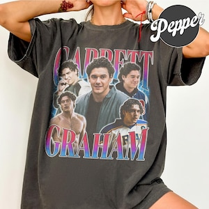 Op de afbeelding: Een antracietgrijs T-shirt met korte mouwen en een grafische print in vintage-stijl van Garrett Graham. Het ontwerp bevat vier verschillende fotografische portretten van dezelfde persoon, met de naam Garrett Graham in grote, dikke letters met kleurverloop.