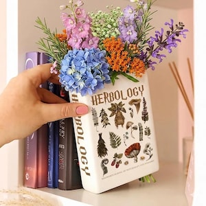 May include: A white ceramic vase shaped like a book, labeled "Herbology," filled with colorful flowers. The vase features botanical illustrations and text. The flowers include blue hydrangeas, purple lavender, and orange accents.