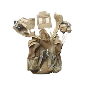 May include: A tan military-style tactical vest with multiple pouches, straps, and attachments. The vest has a modular design with various compartments and pockets for carrying equipment. It is designed for utility and functionality.