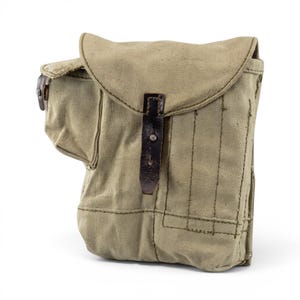Puede incluir: Una bolsa utilitaria de lona color canela con un compartimento principal grande y una solapa redondeada asegurada por una correa de cuero marrón oscuro y un perno metálico. El exterior incluye un bolsillo lateral y paneles cosidos.