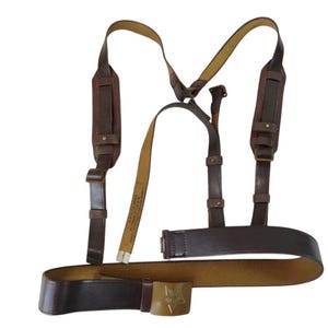 Op de afbeelding: Een bruine leren militaire riem- en bretelset. De bretels hebben schouderbanden en worden aan de riem bevestigd. De riem heeft een goudkleurige gesp met een sterembleem. De set lijkt vintage te zijn.