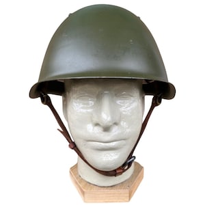 May include: An olive green military helmet with a rounded top and a brim. The helmet is displayed on a mannequin head with brown leather chin straps. The mannequin is mounted on a wooden base.