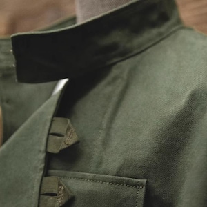 May include: Close-up of a dark green jacket with a structured collar and a small pocket. The jacket has a textured fabric and visible stitching details. The collar is folded down, and the jacket appears to be made of a durable material.