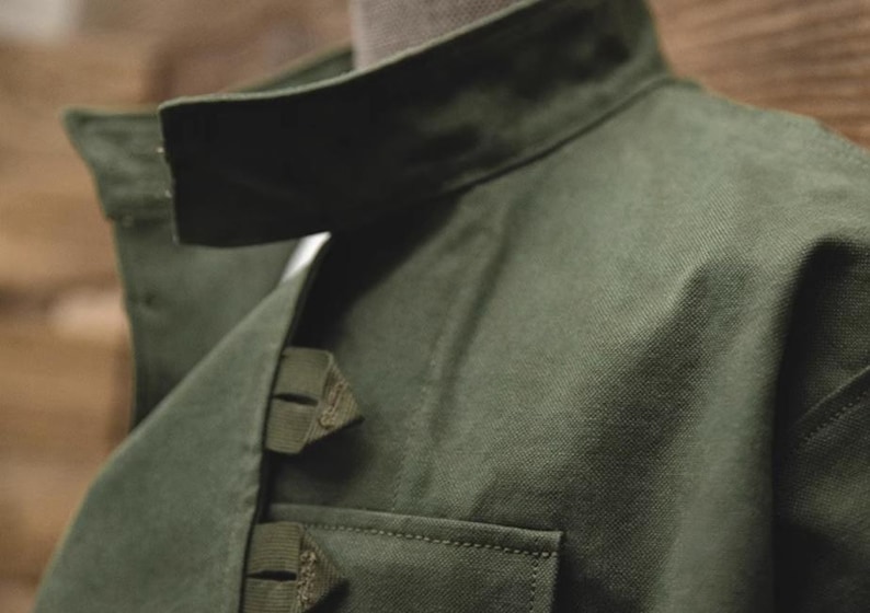 May include: Close-up of a dark green jacket with a structured collar and a small pocket. The jacket has a textured fabric and visible stitching details. The collar is folded down, and the jacket appears to be made of a durable material.