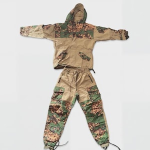 May include: A two-piece camouflage suit consisting of a hooded jacket and matching trousers. The garment features a tan base color with green, brown, and black pixelated camouflage patterns on the hood, shoulders, and lower legs.