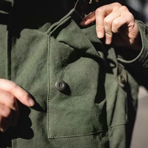 May include: Close-up of a dark green jacket with a buttoned pocket and buttoned cuffs. The jacket has a classic design with visible stitching details. The person's hand is pointing at the pocket.