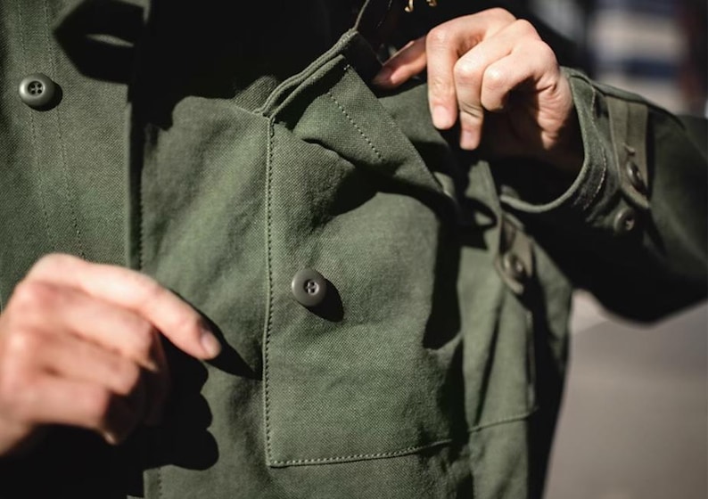 May include: Close-up of a dark green jacket with a buttoned pocket and buttoned cuffs. The jacket has a classic design with visible stitching details. The person's hand is pointing at the pocket.